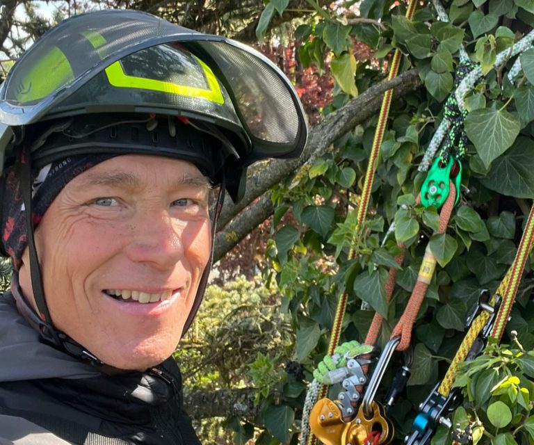 Baumpfleger Patrick Weihe mit Schutzhelm, umgeben von grünen Pflanzen und Kletterausrüstung.